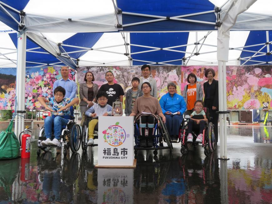 青色と白色の屋根のテントの下で、5人の車いすに乗った方々と6人の男女が市長と並んで立った記念写真