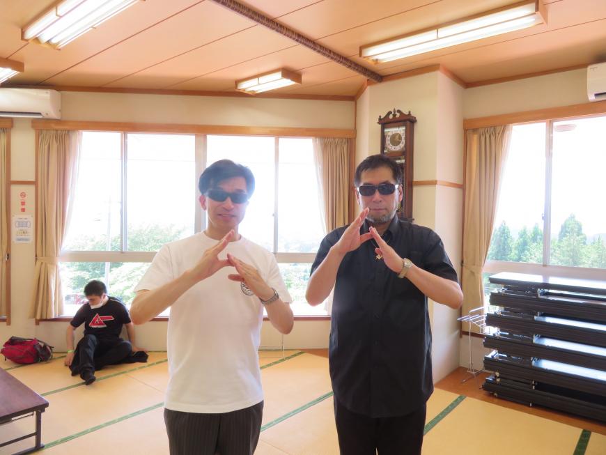 畳の部屋でサングラスをかけた市長と男性が両手でカタカナのムを作るようなポーズをしている写真