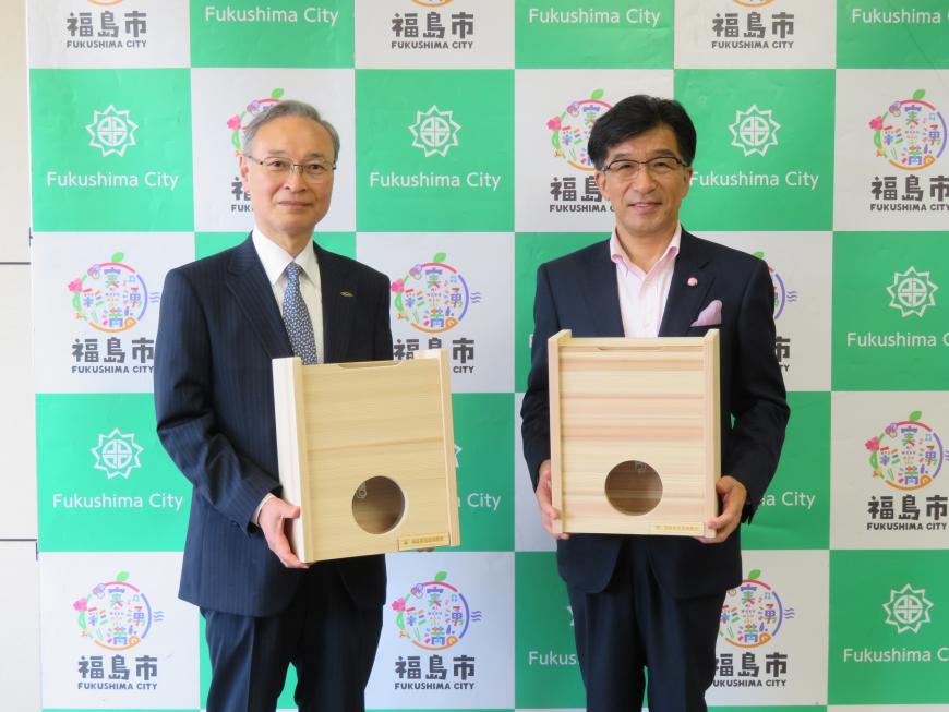 バックパネルの前で市長と男性がそれぞれ木製の募金箱を持って立っている写真
