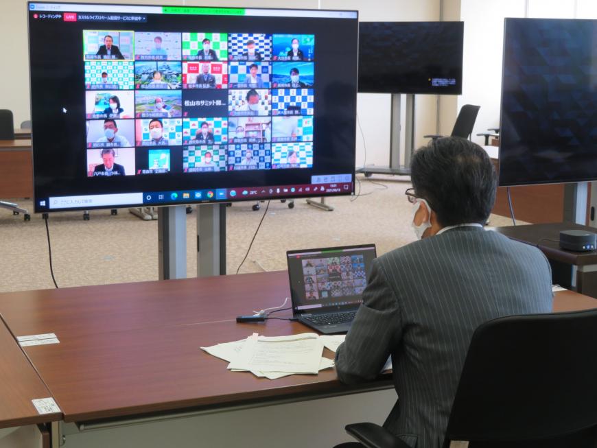 ノートパソコンが置かれたテーブルの席に座った市長が、テーブルの前に設置された、リモート参加者たちの画像が映し出された大型モニターを見ながら会議を行っている写真