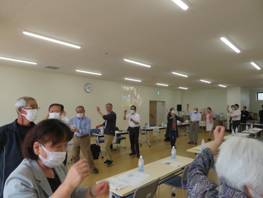 長机が並んでいる会場で、参加者が円形になって踊りをしている写真