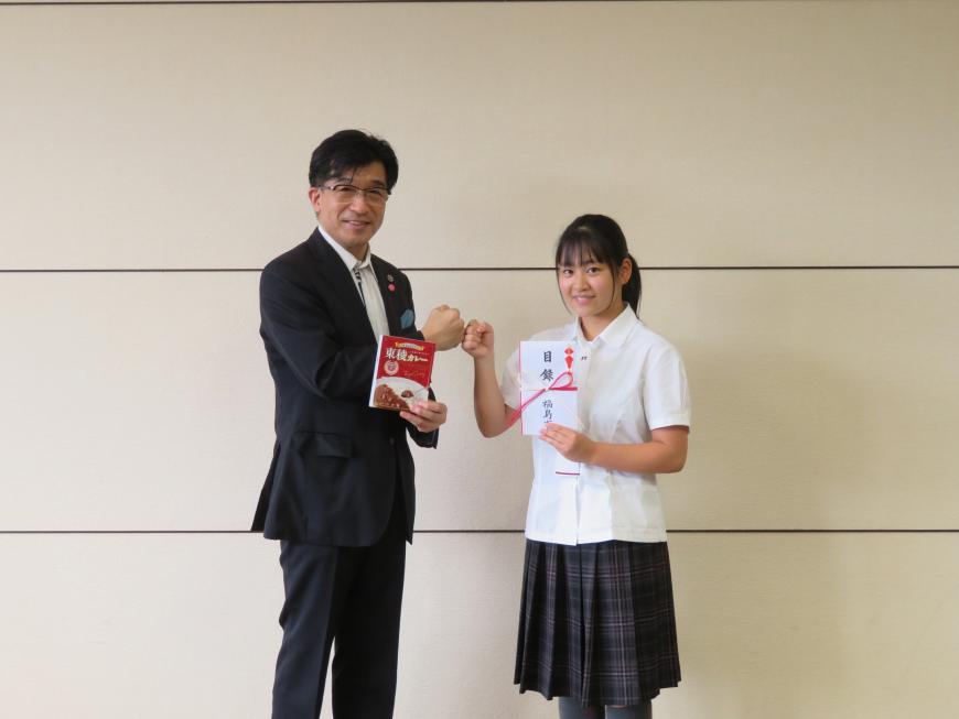 壁の前で、目録封筒を持った制服姿の一条さんと、カレーの箱を持った市長が笑顔でグータッチをしている写真