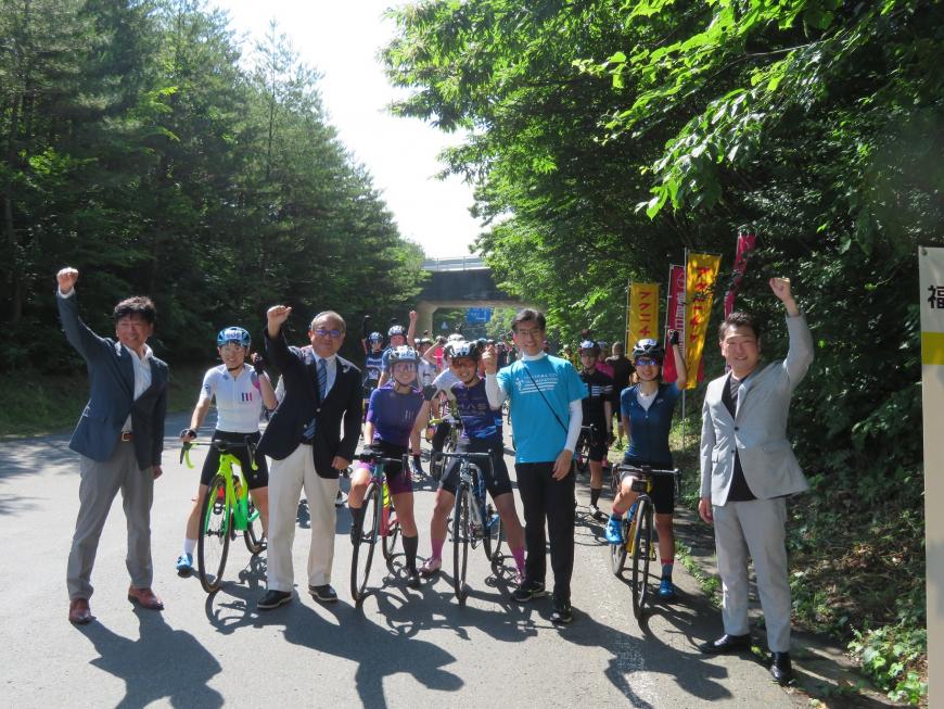 自転車にまたがって集まっている参加者の前に、市長の含む男性4名が立ち、参加者みんなでガッツポーズをしている写真