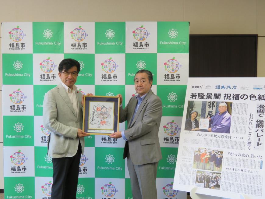 福島市のバックパネルの前で、市長と男性が額に入ったサイン入りのイラストの色紙を持って立ち、右側に大きく拡大した福島民友新聞の「若隆景関祝福の色紙」の記事が飾られている写真