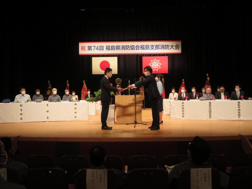 多くの来賓の方が座っているステージで、市長が男性に賞状を渡している写真