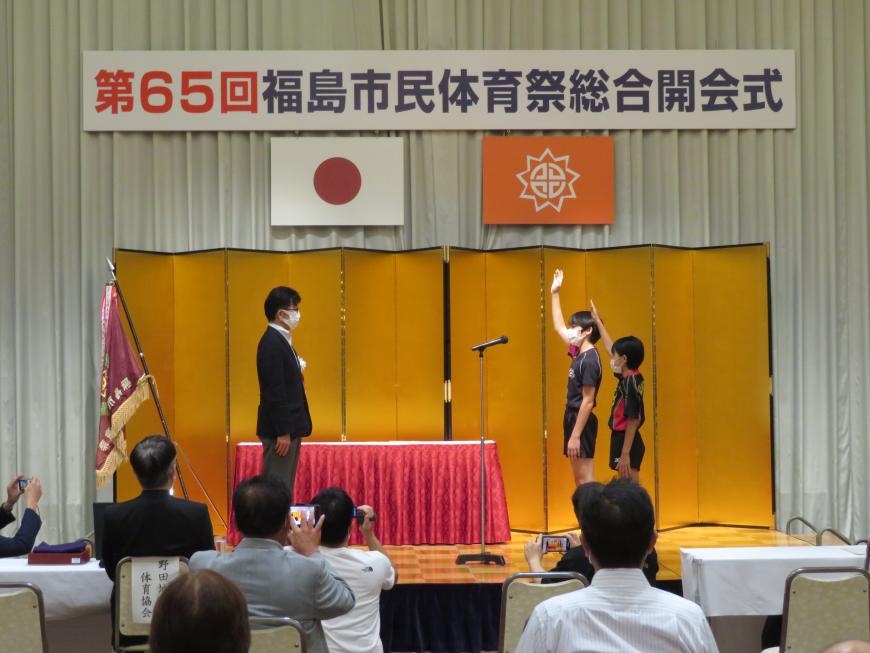 「第65回福島市体育祭総合開会式」と書かれた舞台看板の下に日本国旗と福島市の市章が吊るされ、金屏風が立てられた舞台で、市長と向かい合って立った2人の児童が片手をあげ選手宣誓をしている写真