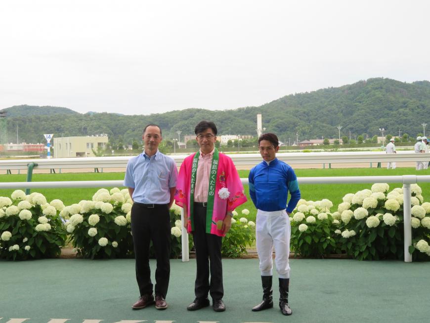 競馬場のコース外で、法被を着た市長と騎手と男性が並んでいる写真
