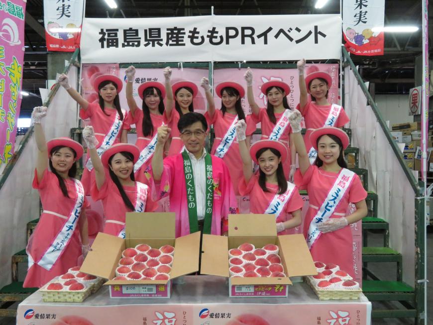 箱詰めされた桃が置かれた後ろで、お揃いのピンクの帽子、ワンピース姿の10人のミスピーチが市長を中心としてひな壇状に2列に並んで右手を突き上げている写真