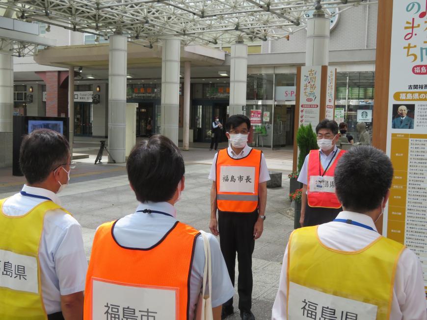 駅前で蛍光のオレンジや黄色、ピンクのビブスを着用した男性達が「福島市長」と胸に書かれたオレンジ色のビブスを着た市長と向かい合って立っている写真