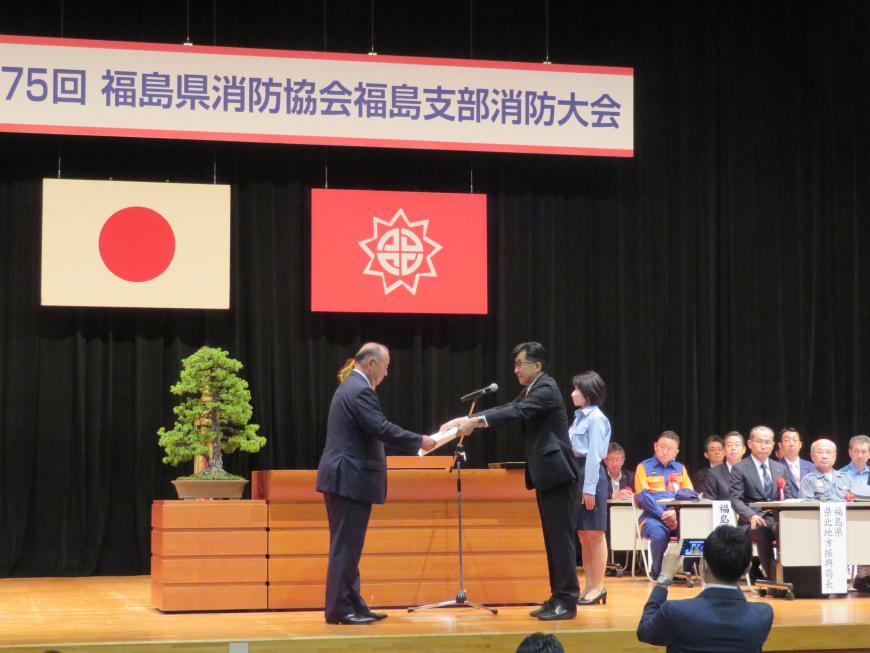 ステージに多くの来賓の方が座っている中、演台前で市長が男性に賞状を渡している写真