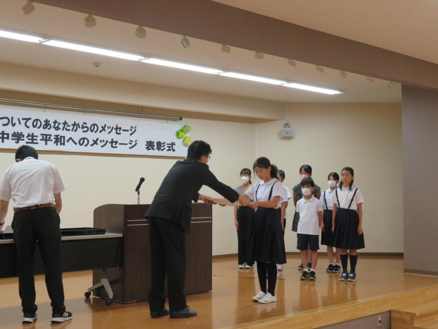 入賞した生徒、児童がステージで整列している前で、市長が女子児童に賞状を渡している写真