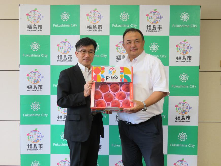 バックパネルの前で、市長と男性でFBOXと書かれた箱を持っている写真