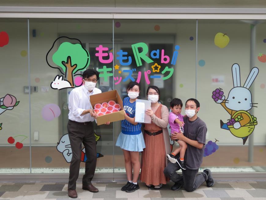 「ももRabiキッズパーク」と書かれたガラス戸の前で市長と親子4人が並んで、市長と女の子で桃が入った箱を持っている写真