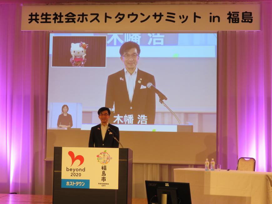 市長が演台に立っている様子が後ろのスクリーンに映し出されている写真