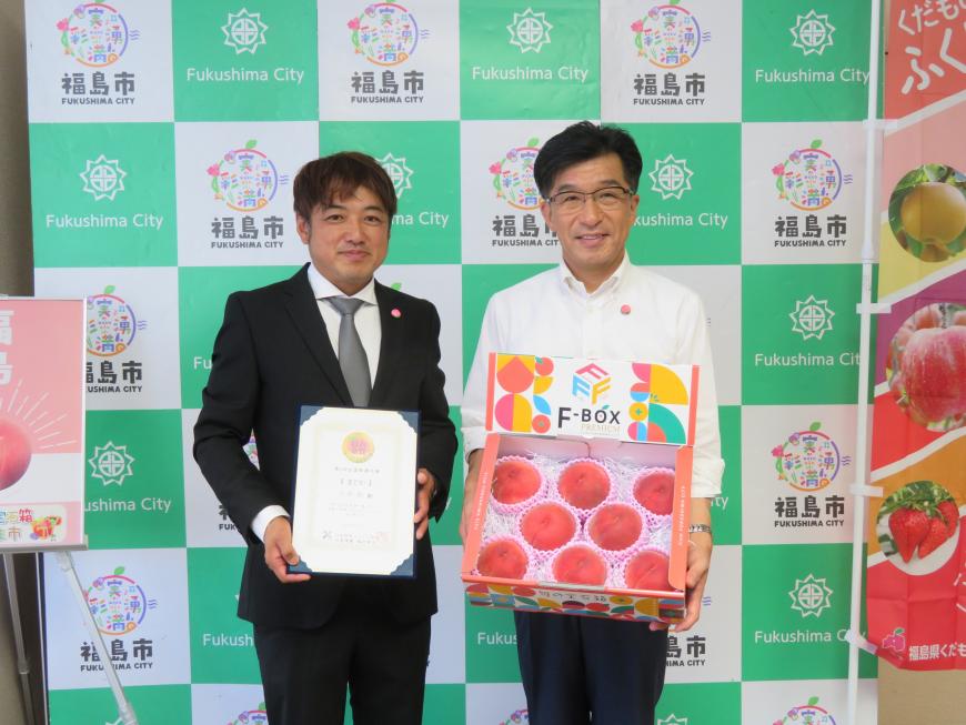 バックパネルの前で、市長が桃が入った箱を持ち、横の男性が賞状を持って立っている写真