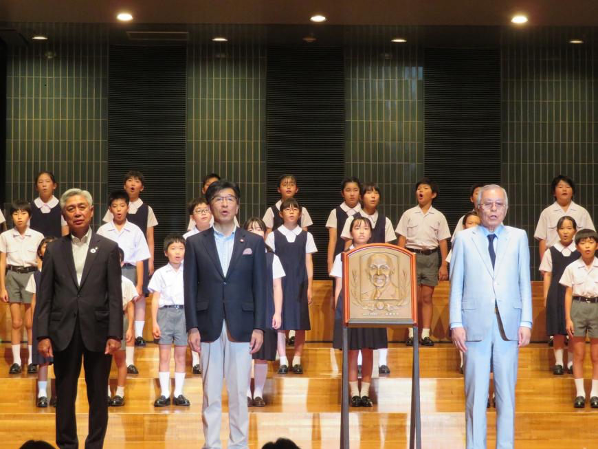銅板プレートの横に市長と男性2名が立ち、後方には児童たちがひな壇状に並んで合唱している写真