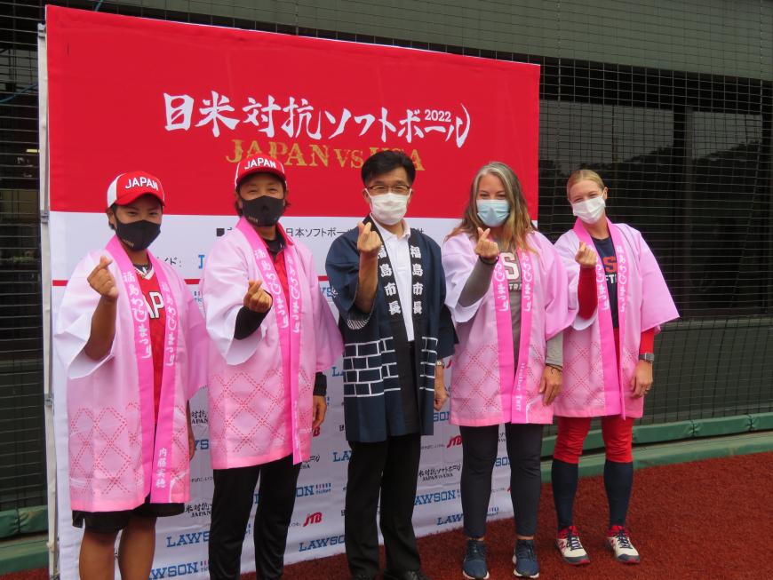 日米対抗ソフトボールの看板の前で同じピンクの法被を着た日本チーム2人とアメリカチーム2人の間に紺色の法被を着た市長が立ち、指ピースのポーズしている写真