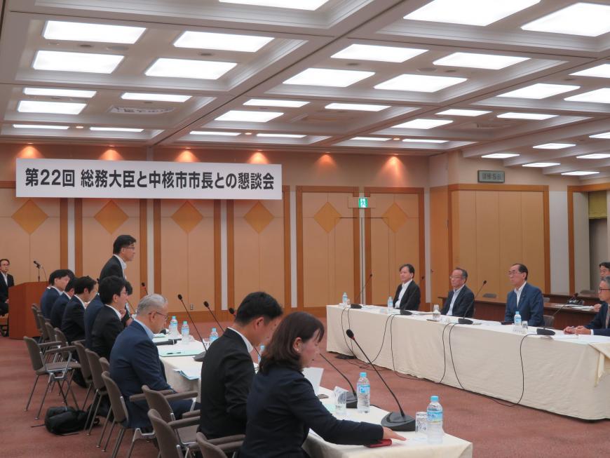 「総務大臣と中核市市長との懇談会」と書かれた部屋で机を対面するように並べられて座り、市長が自席で立って意見を述べている写真