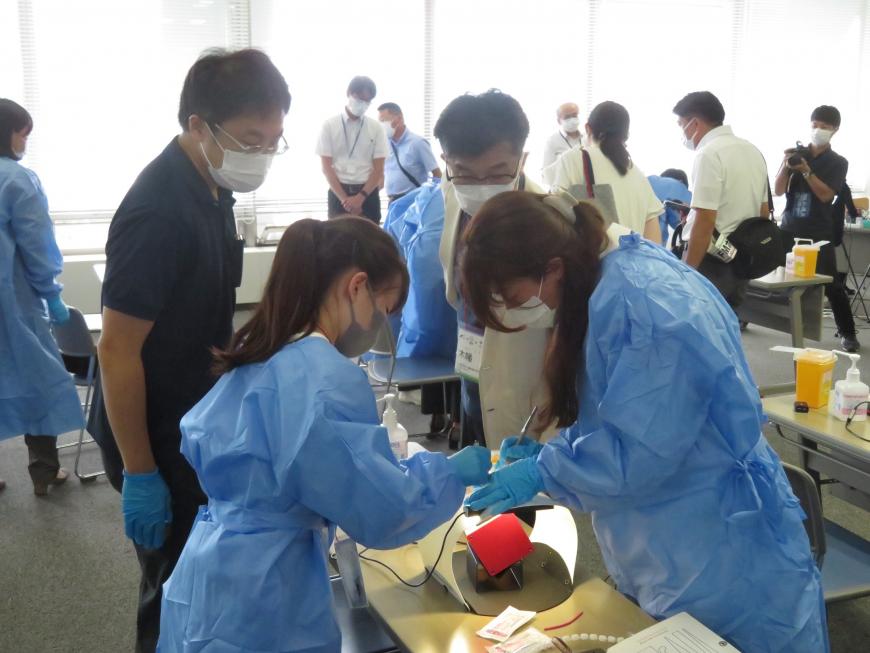 青の作業着を着て訓練している女性2人を市長が見ている写真