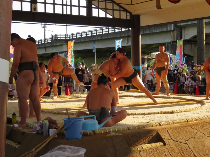 多くの見学者が見ている中、土俵で黒のまわしをつけた力士と白のまわしをつけた子供が稽古をしている写真
