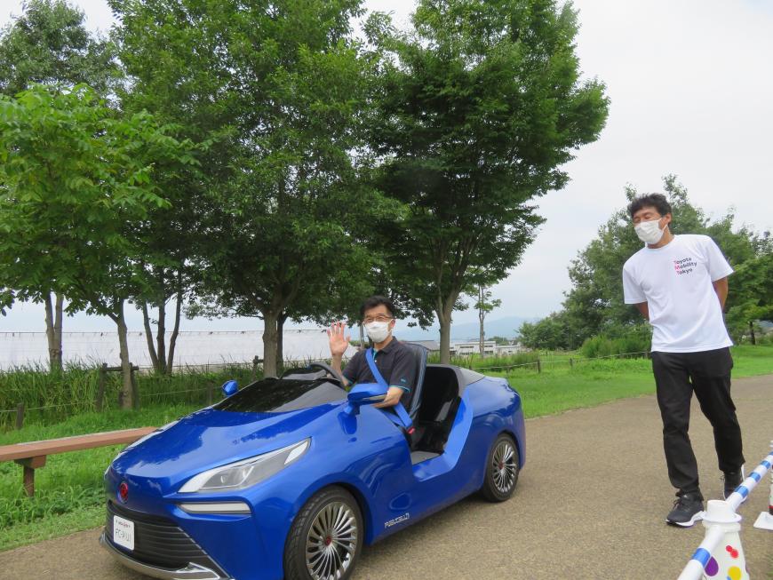 男性が見ている中、公園内の道路を市長が青のオープンカーに乗り、右手を挙げている写真
