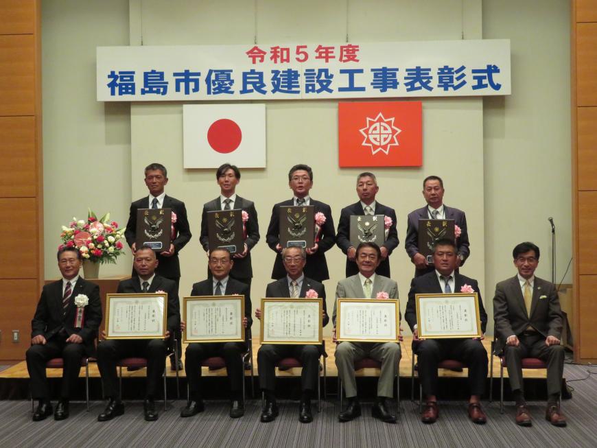 前列に額入りの賞状を持った男性5名が座り、後列に盾を持った男性5名が立っている写真