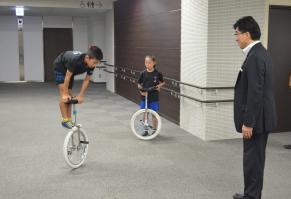 部屋の一室で、一輪車の車輪の上でバランスを取りながらで静止する小学生の姿を見つめている市長の写真