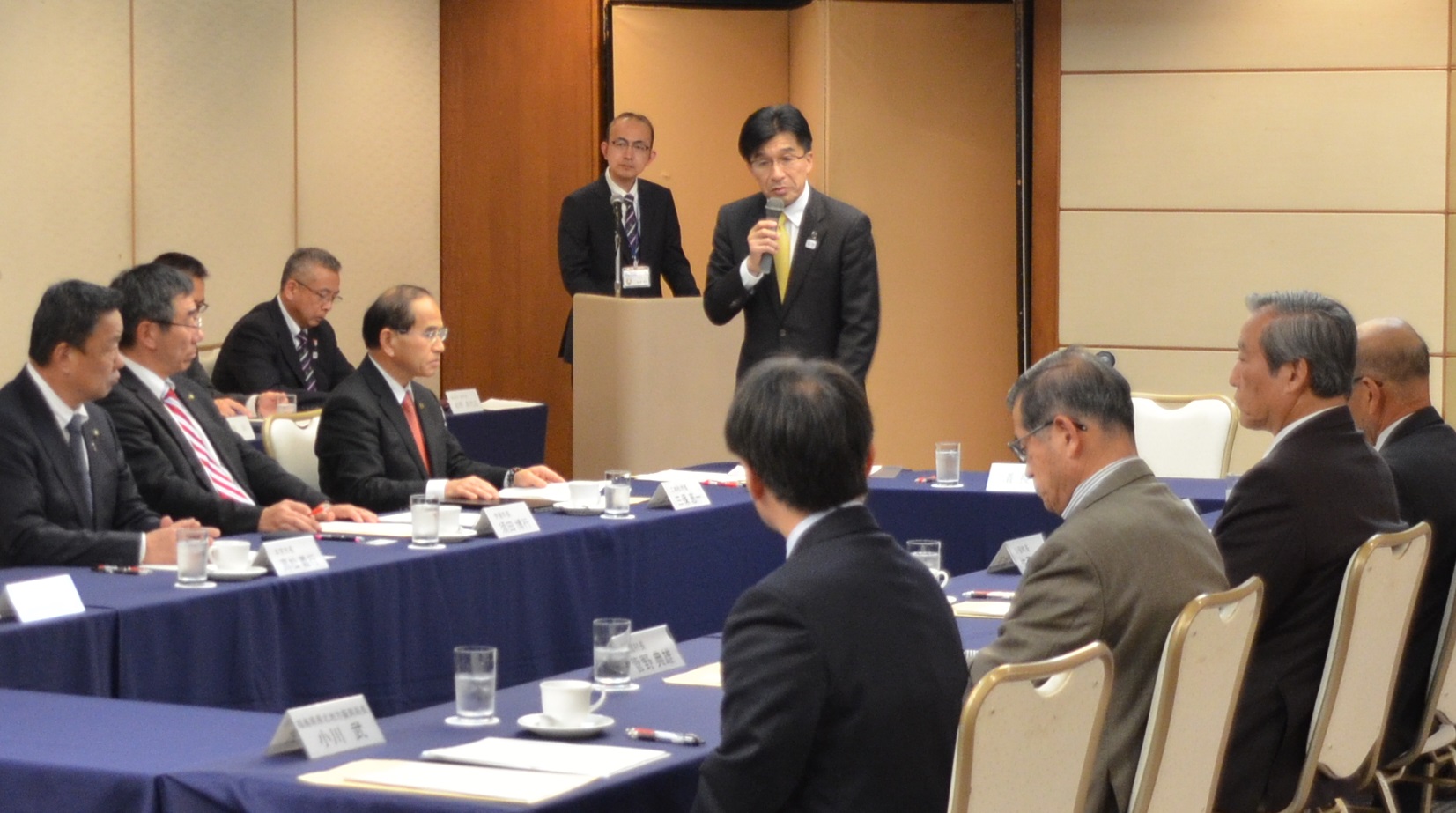 11市町村長がロの字型に設置された席に座っており、市長が席を立ち、マイクを手に持って発言している写真