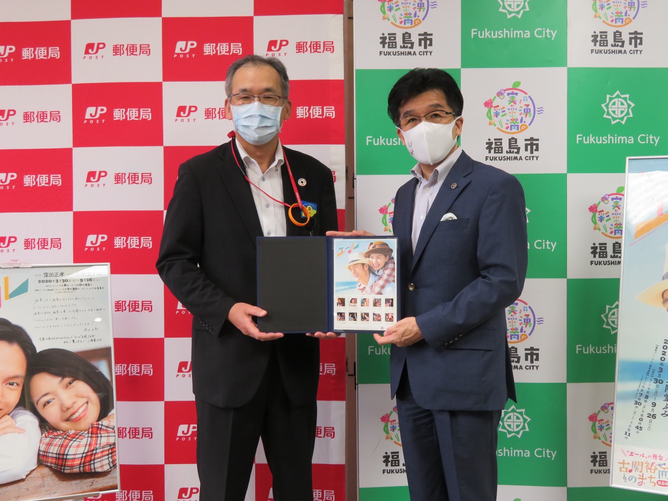郵便局と福島市のバックボードの前で市長と男性が連続テレビ小説エールの主人公の2人や、ドラマのシーンが描かれたオリジナル切手が入ったファイルを一緒に持って記念撮影している写真