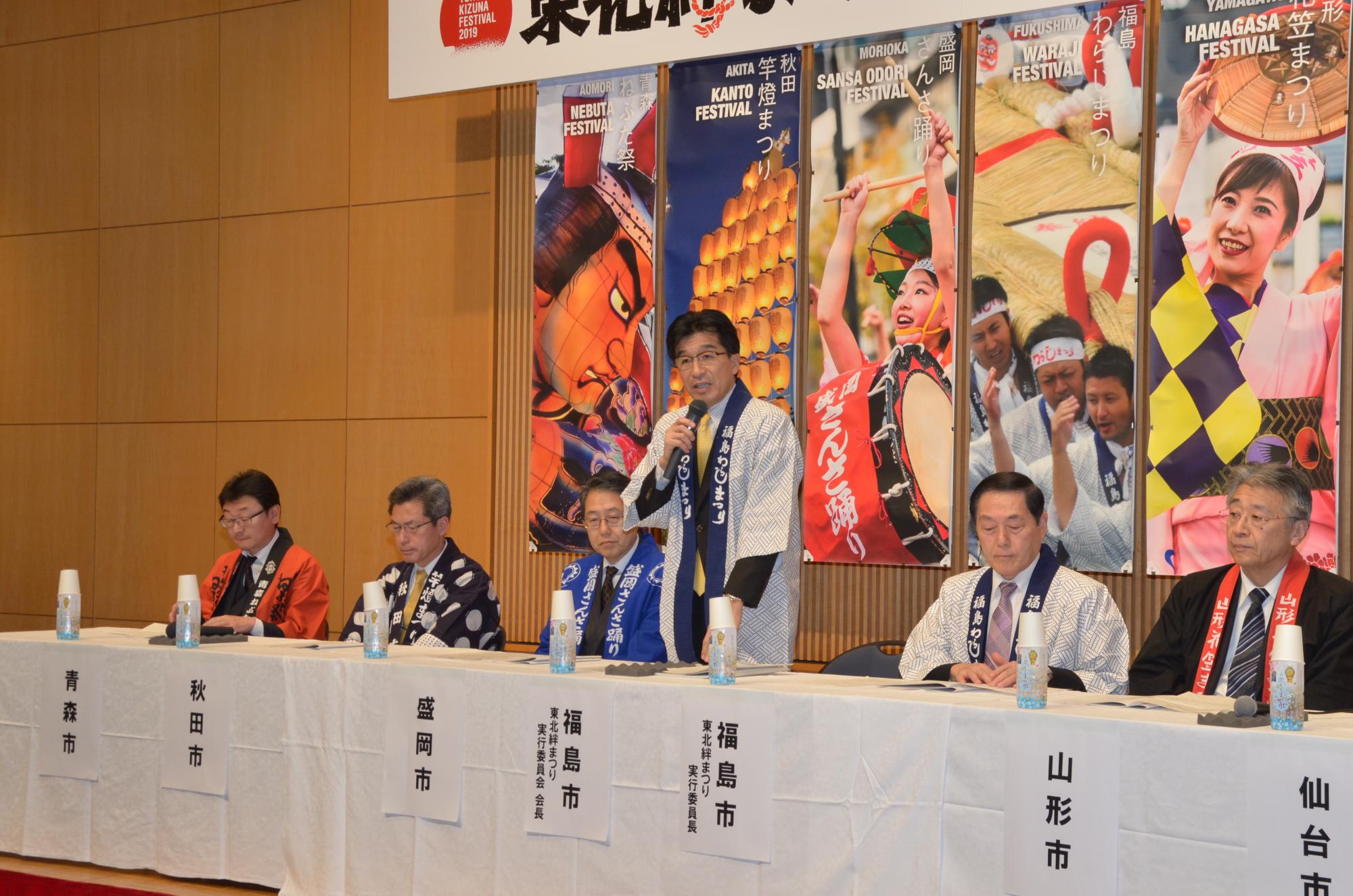 はっぴ姿の市長がマイクを手に持ち、立って話をしており、ほかの関係者は席に座り話を聞いている写真