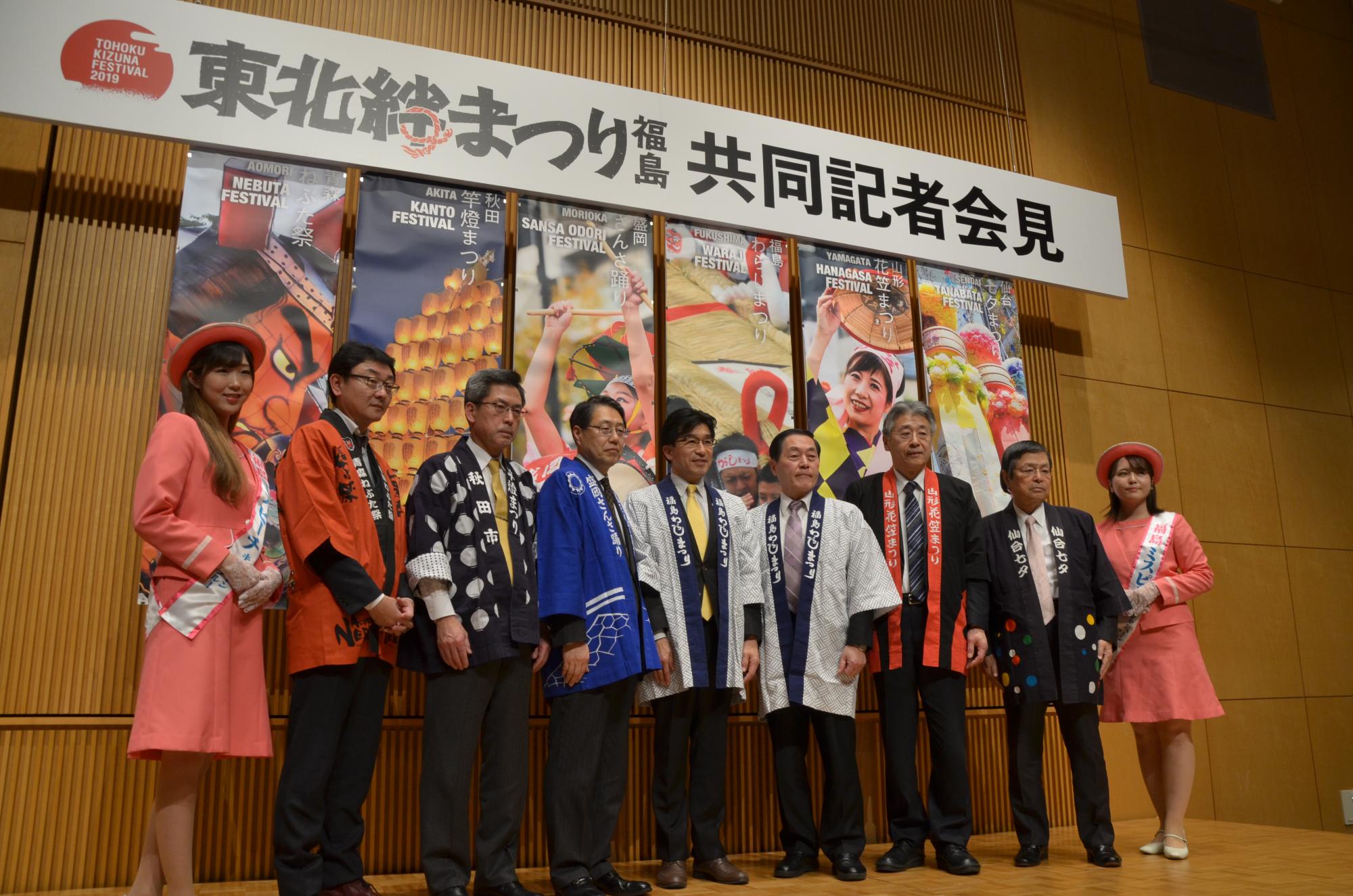 「東北絆まつり福島共同記者会見」の吊りパネルの下、カラフルなはっぴ姿の各市長達とミスピーチキャンペーンの女性が並んでいる写真