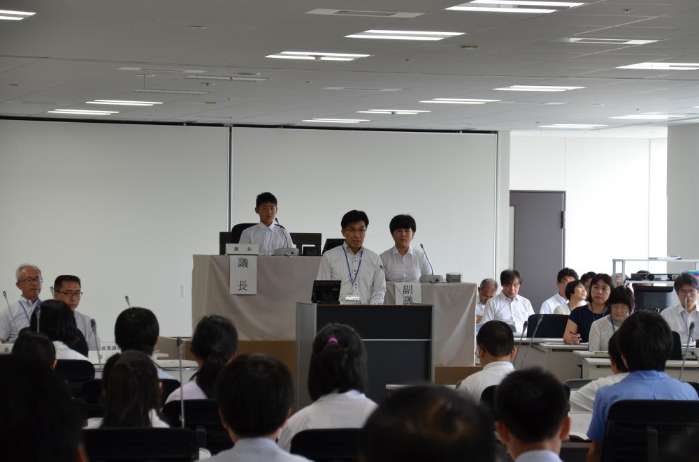 会議室で議会を模して配置された席に児童など大勢の出席者が座る中、議長席の前に置かれた演台に市長が立っている様子の写真