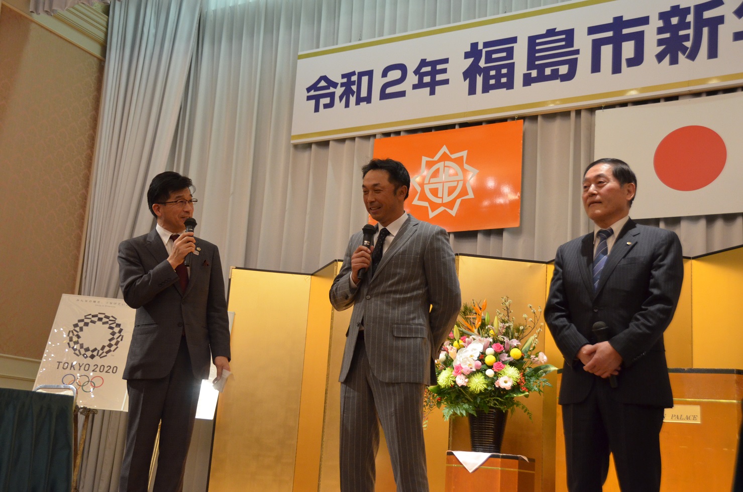 「令和2年福島市新年市民交歓会」と書かれた舞台看板が掲げられたステージ上に立つ関係者の男性と宮本慎也さんに向かって市長がマイクを持ち話をしている写真