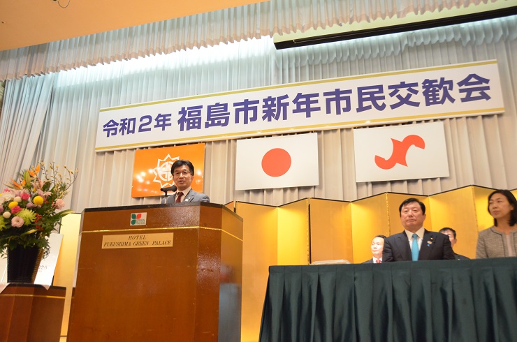 「令和2年福島市新年市民交歓会」と書かれた舞台看板が掲げられたステージ上に関係者が座り、中央の檀上で挨拶を行っている市長の写真