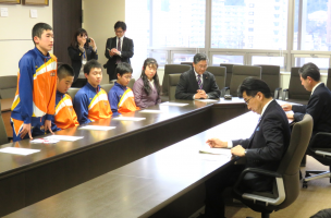 市長応接室で、岡山スポーツ少年団バレーボール部の子供たちが並んで席に座っており、男の子が席を立ち、市長へ話をしている写真