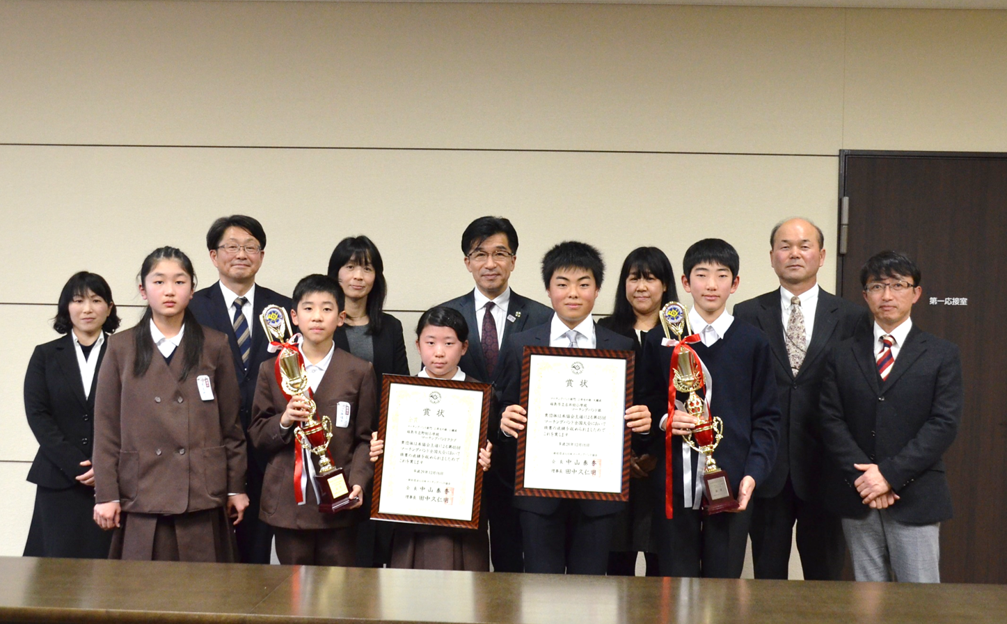 賞状、トロフィーを手に持った制服姿の小学生達と市長と関係者が並んでいる写真