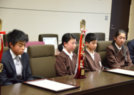 応接室の席に小学生達が座り、机の上にトロフィーと賞状が置かれている写真