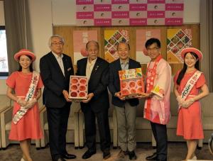 ピンクの帽子を被り、同色のワンピースを着た2名の女性の間に法被を着た市長を含めて男性4名が立ち、桃が入った箱を2名で1箱持っている写真