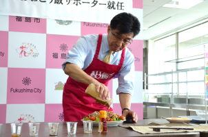 エプロン姿の市長が、お皿に盛られたサラダにドレッシングをかけている写真