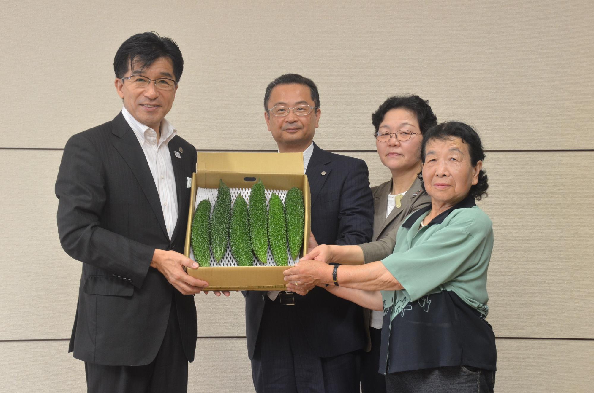 左手に市長、右手にスーツの男性1名と女性2人が並んで立ち、6本の大きなニガウリが入った箱を全員で持って写った記念写真