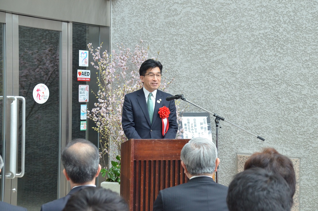 ふくしん夢の音楽堂の玄関前で左胸に赤い胸章リボンをつけた市長が演壇に立ちマイクの前で話をしている写真