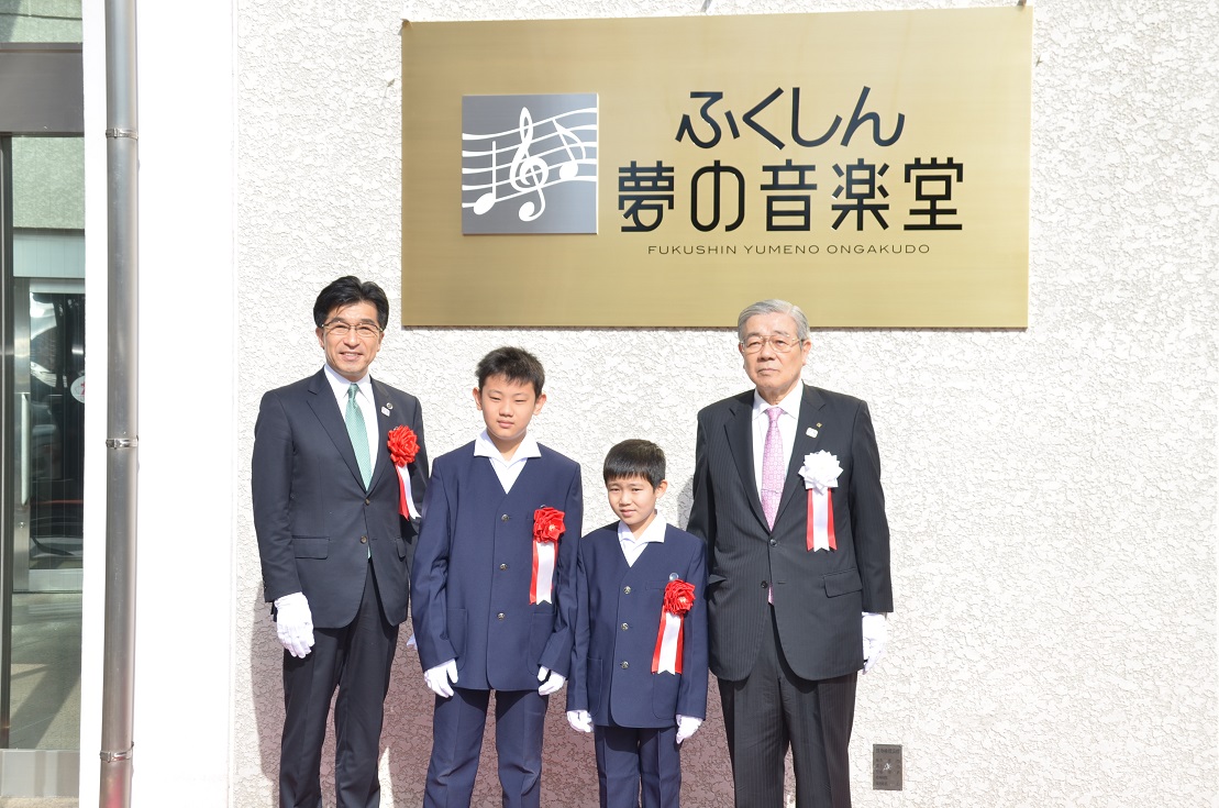 「ふくしん夢の音楽堂」と書かれた金色の看板の前で左胸に胸章リボンをつけている市長と男の子2人と男性が横並びで記念撮影をしている写真