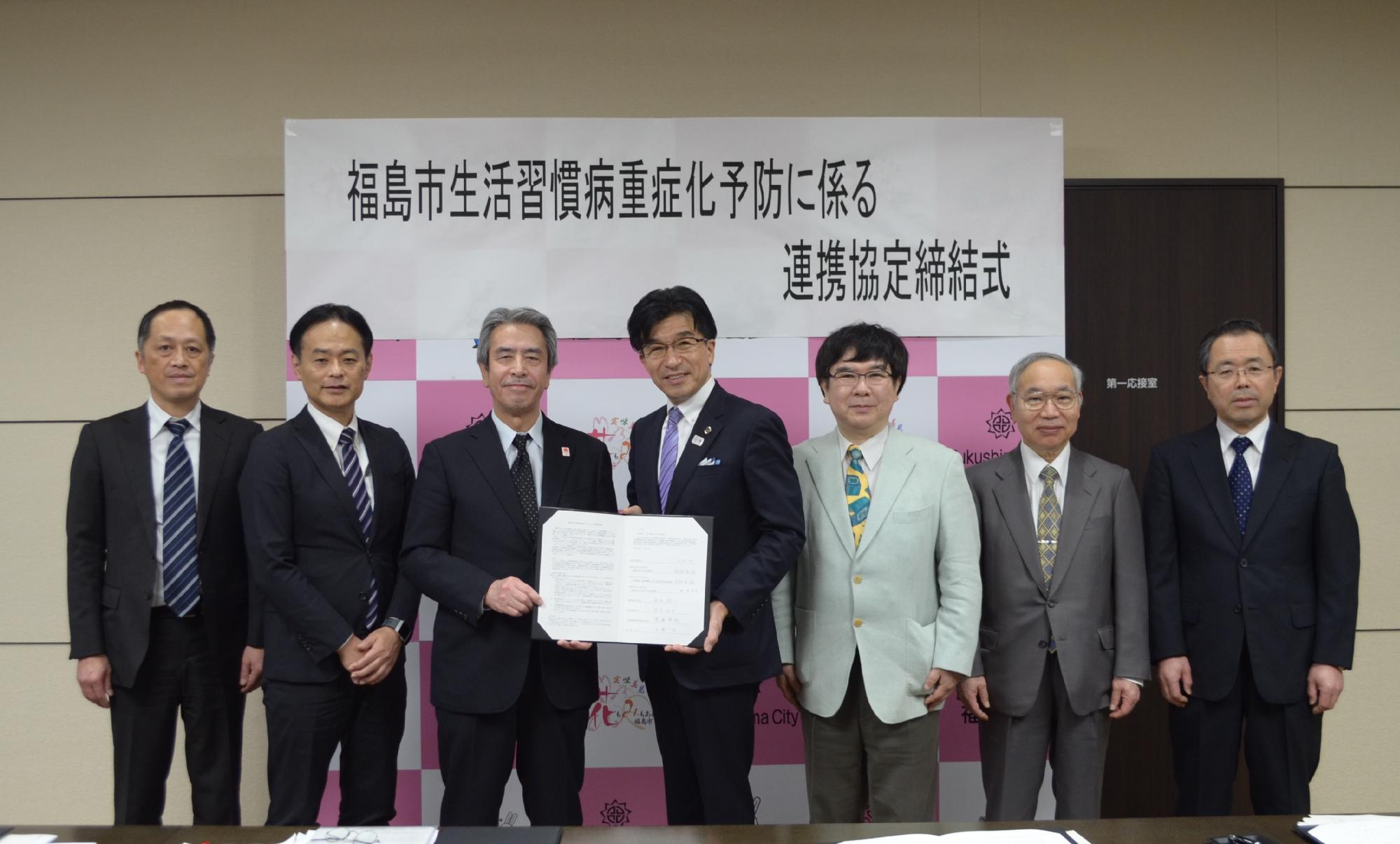 バックパネルに「福島市生活習慣病重症化予防に係る連携協定締結式」の横断幕が掲げられた前で、市長と男性が開いた締結書を手に持ち、他の関係者達と一緒に並んでいる写真