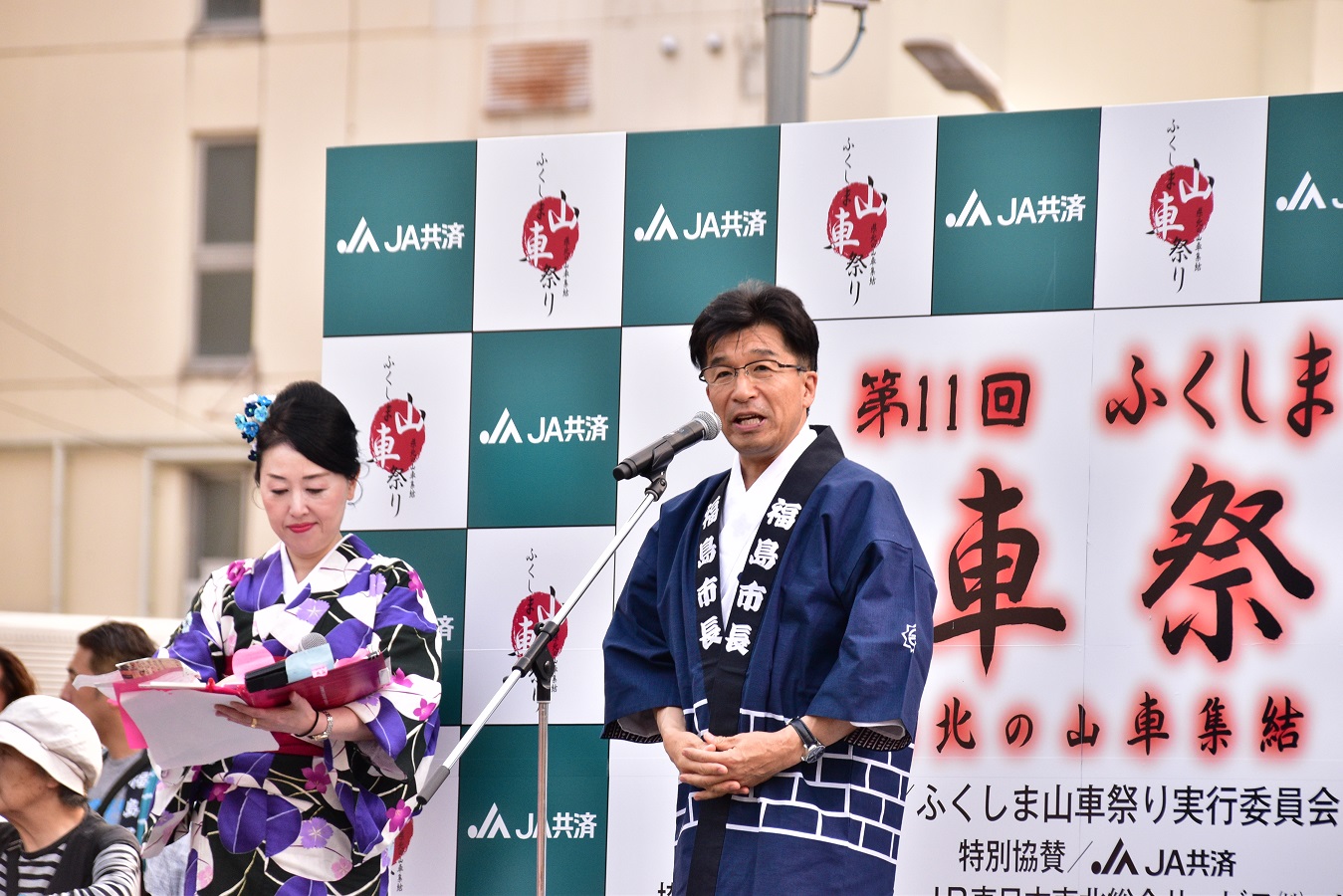 着物姿の女性が立っている横で、はっぴ姿の市長がマイクスタンドの前で話をしている写真