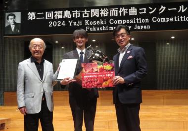 「第二回福島市古関裕而作曲コンクール」と書かれた横断幕の前で、トロフィーを抱えた男性の両脇にフルーツのパンフレットを持った市長と賞状をもった男性が立っている写真