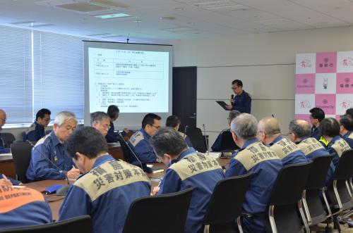 大きなスクリーンにスライドが映し出された会議室で、災害時のユニフォームを着た出席者たちが長机に向かい合って座っている写真