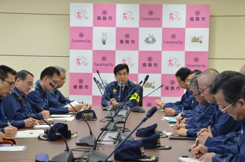 福島市のバックパネルの前に会議用机の中央に複数のマイクが並び、中央に市長と両端に紺色のユニフォームを着た男性たちが座っている写真