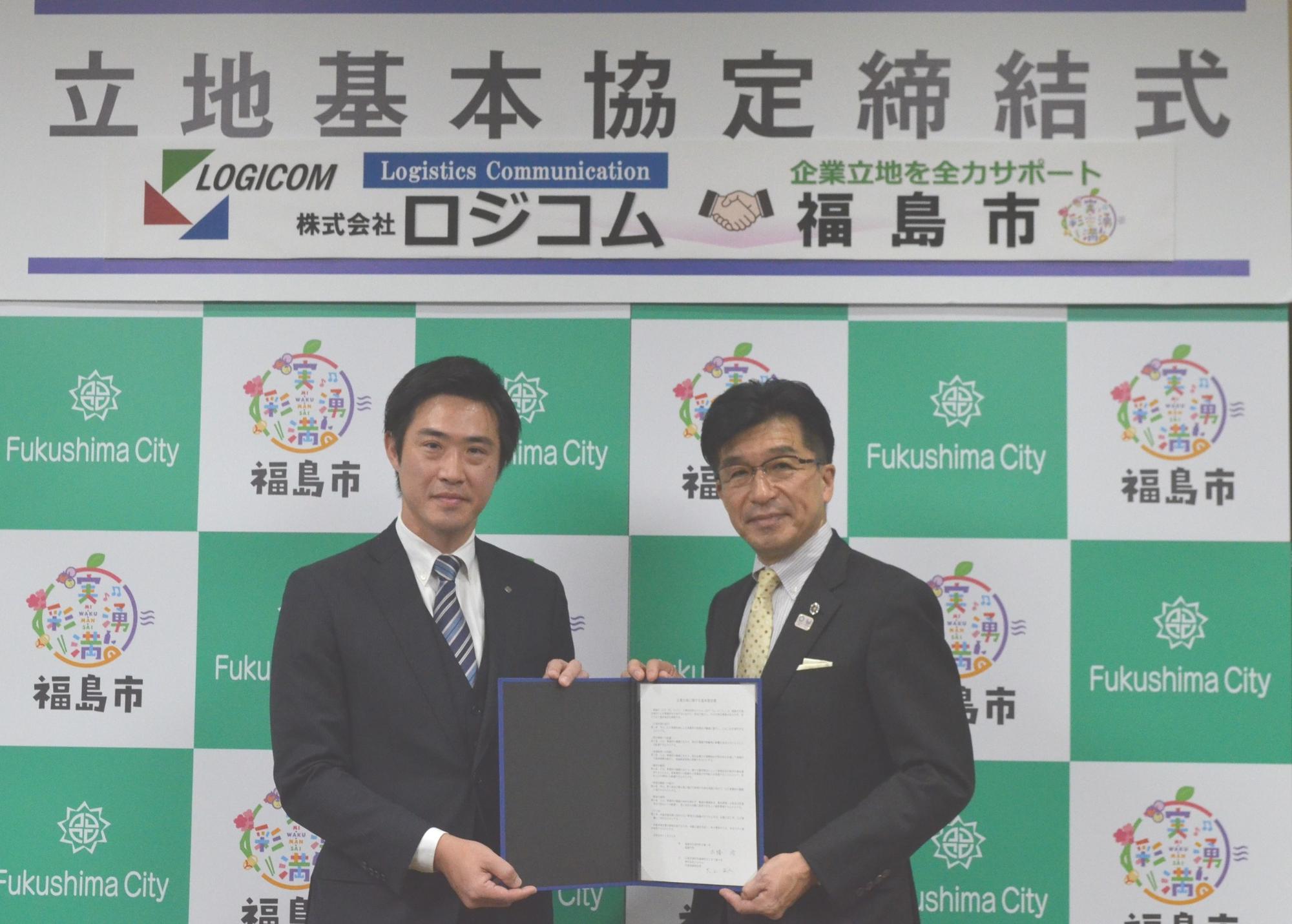 福島市のバックボードの上に株式会社ロジコムと福島市のロゴと「立地基本協定締結式」と書かれたパネルの前で協定書を持った市長と男性が記念撮影している写真