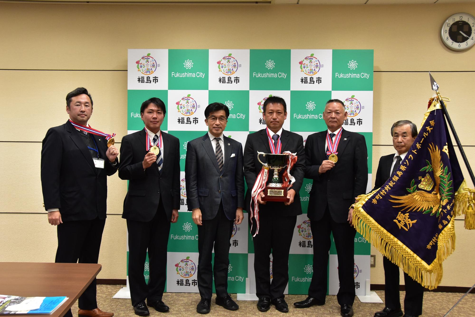 福島市のバックパネル前で、市長とソフトボール大会優勝旗とトロフィーを手に持ち、首にメダルを掛けた福島市チームのと関係者5人が並んでいる写真