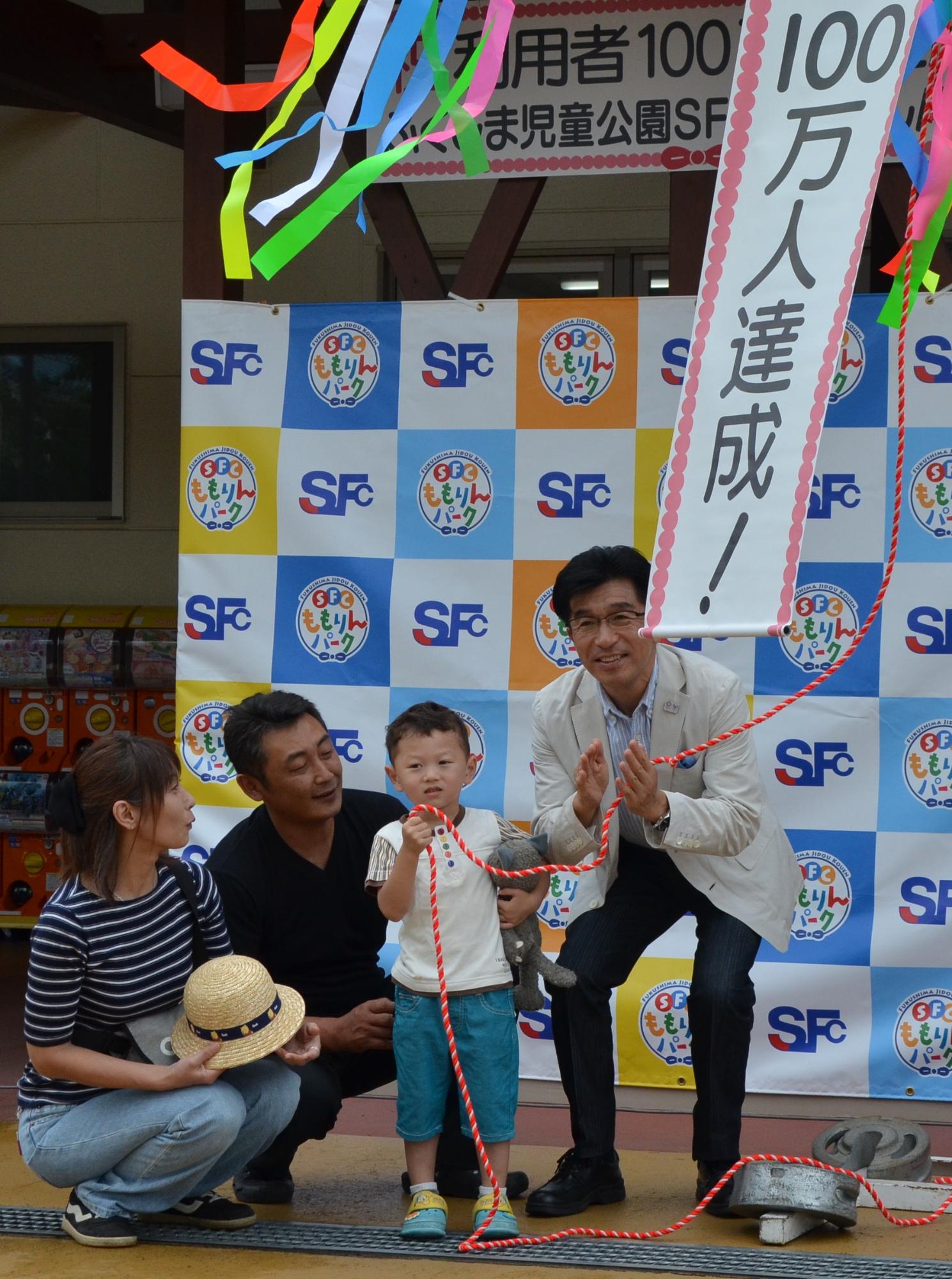 「SFCももりんパーク」のバックボードの前で、くす玉の赤い紐を手に持った小さな男の子の左側に両親がしゃがみ、男の子の右で市長が赤い紐を持ち腰を落としながら拍手をしている頭上に、くす玉の飾りと「100万人達成!」の垂れ幕が見える写真